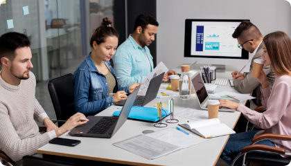 A diverse group of four young professionals working together at a table with laptops, documents, and a large monitor displaying graphs and charts in a modern office.