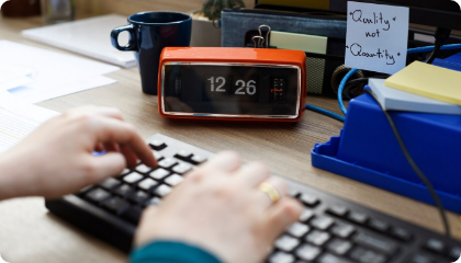 Close-up of hands typing on a computer keyboard with a retro-style clock and a sticky note that reads “Quality not Quantity” in the background.
