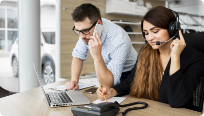 Two coworkers in an office, one speaking on a phone while the other wearing a headset writes notes, with laptops and office supplies on the desk.