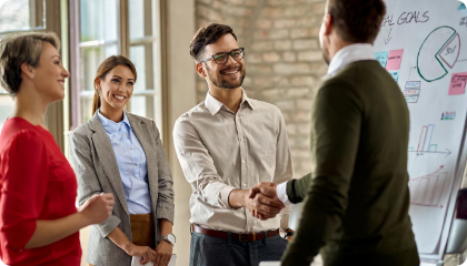A business meeting with smiling professionals shaking hands, with two women standing nearby and a whiteboard with charts and graphs in the background.